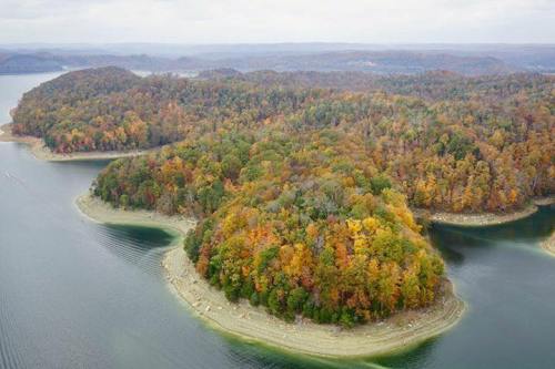 Aerial view of a colorful forested island surrounded by calm water, showcasing autumn foliage and distant hills.