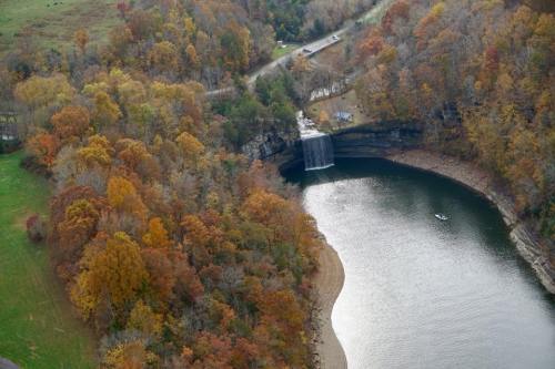Aerial view of a serene river with a waterfall, surrounded by autumn foliage and a winding road.