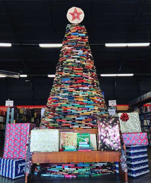 A creative Christmas tree made of stacked books, surrounded by colorful wrapped presents in a library setting.