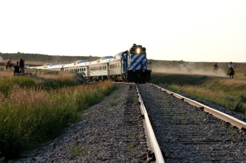 A train travels along a dusty track, with grasslands and distant riders in the background under a clear sky.