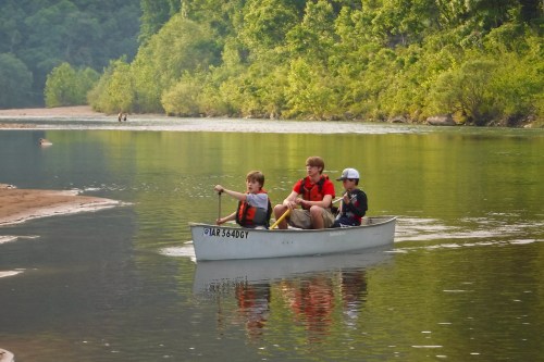 Three children in a canoe paddle on a calm river surrounded by lush greenery.