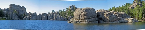A panoramic view of a serene lake surrounded by large rock formations and lush green trees under a clear blue sky.