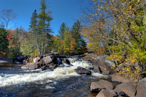 A serene river flows over rocks, surrounded by colorful autumn trees under a clear blue sky.