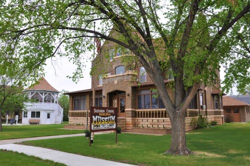 Historic brick building with a sign reading "Stutsman County Museum," surrounded by green grass and trees.