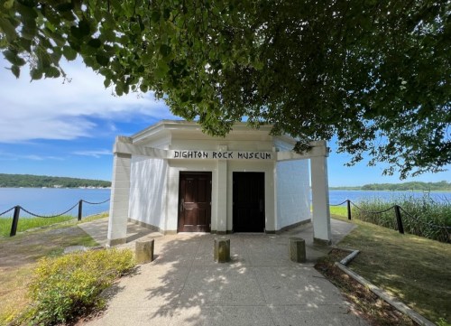 Entrance to Dighton Rock Museum, surrounded by greenery and overlooking a body of water under a blue sky.