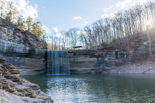 A serene waterfall cascades into a calm lake, surrounded by rocky cliffs and bare trees under a clear blue sky.