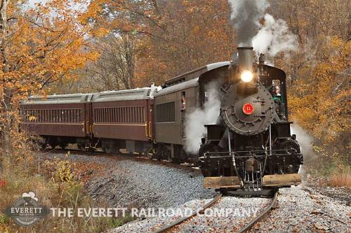 A vintage steam train chugs along a scenic track surrounded by autumn foliage.