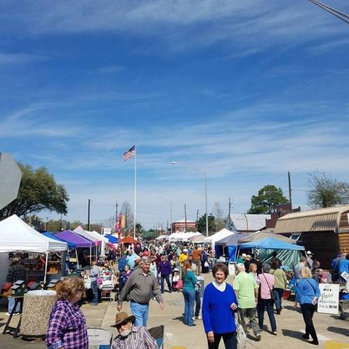 A bustling street market with vendors and crowds under a clear blue sky, featuring tents and an American flag in the background.