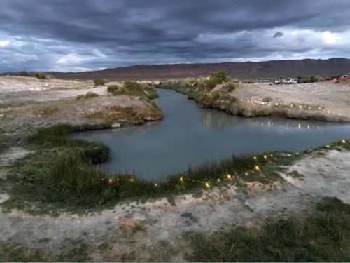 A winding river flows through a barren landscape under a cloudy sky, with lights lining the water's edge.