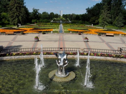 A fountain with a sculpture in a garden, surrounded by colorful flower beds and a pathway leading to the horizon.