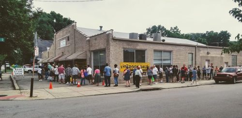 A long line of people waiting outside a building for a book sale, with a sign that reads "Book Sale Open."