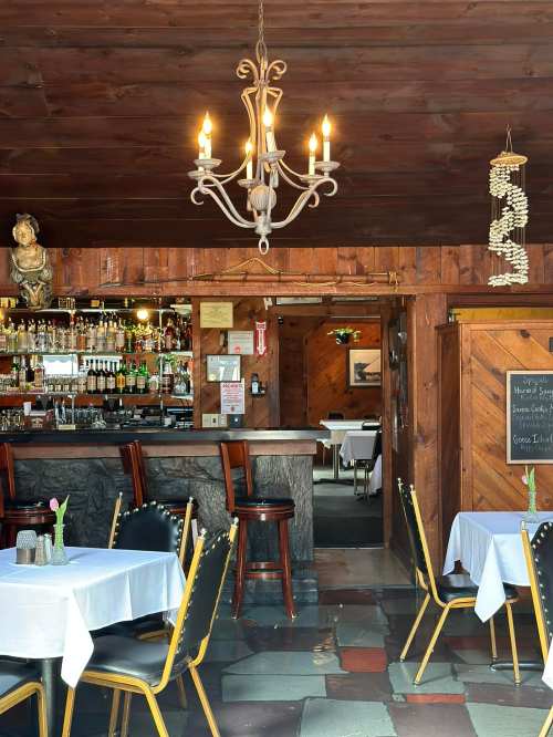 Cozy restaurant interior with a chandelier, bar area, and tables set with white tablecloths and flowers.
