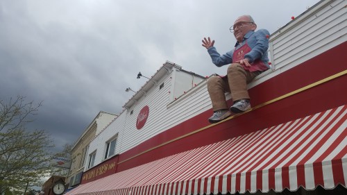 A large, whimsical figure of an elderly man sits on the roof of a shop with red and white striped awning.
