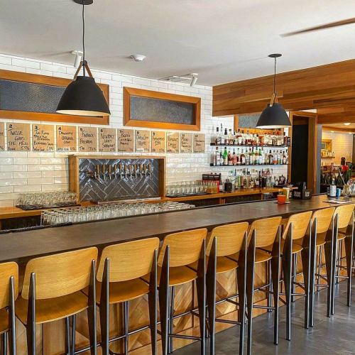 A modern bar with wooden stools, a sleek counter, and shelves lined with various bottles and a menu on the wall.