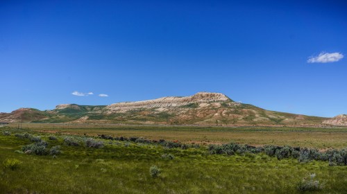 A panoramic view of a grassy landscape with a prominent, layered mountain under a clear blue sky.