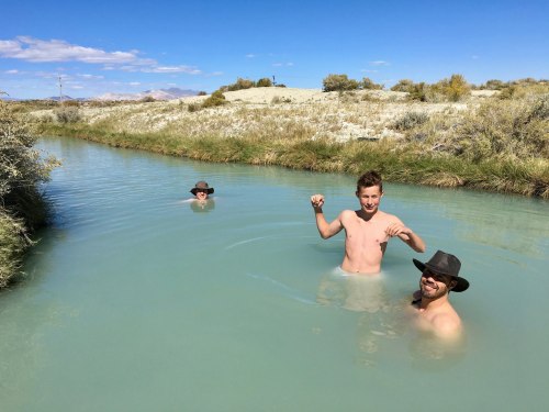 Three boys play in a shallow, light blue stream surrounded by grassy banks and a clear blue sky.