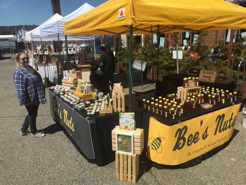 A vendor stands at a market stall with honey jars and wooden crates, under a yellow canopy, surrounded by other booths.