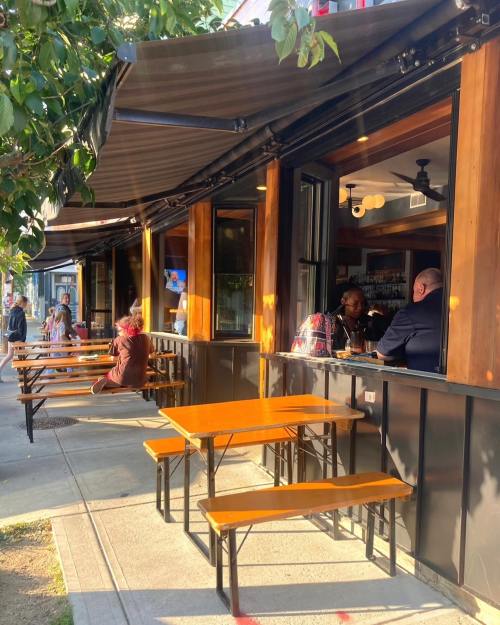Outdoor seating area of a restaurant with wooden tables, people dining, and large windows under a canopy.