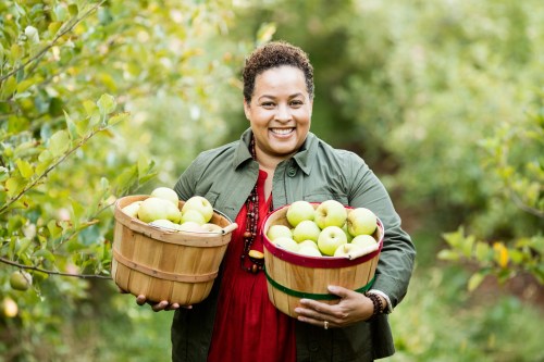 A smiling woman in a green jacket holds two baskets of green apples in an orchard.