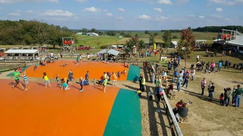 A vibrant outdoor play area with children jumping on a large orange surface, surrounded by families and green fields.