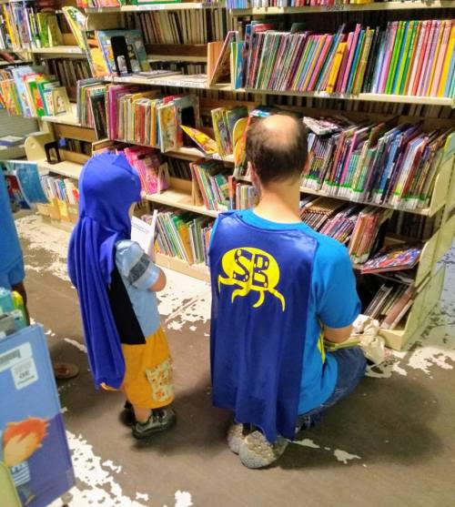 A child in a cape stands beside an adult in a superhero cape, both browsing books in a library.