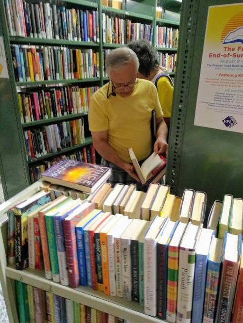 A man in a yellow shirt browses books on a shelf in a library, with another person visible in the background.