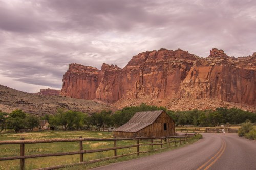A rustic wooden barn beside a winding road, with towering red rock formations and cloudy skies in the background.