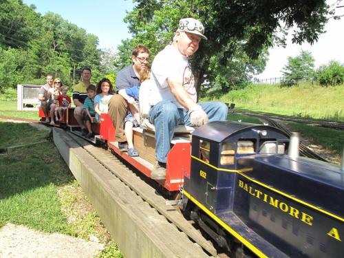 A miniature train with passengers rides along a track in a green park setting on a sunny day.