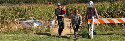 A family walks through a cornfield, with a scarecrow nearby and a barrier at the entrance to the path.