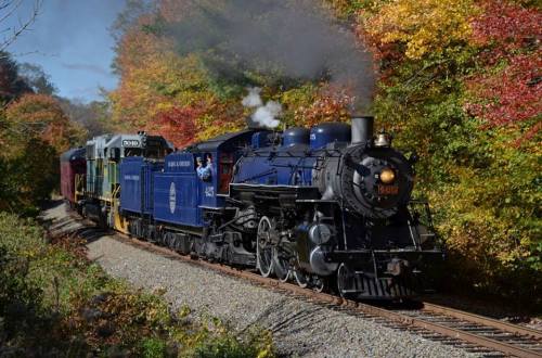 A vintage steam locomotive travels along a scenic railway surrounded by colorful autumn foliage.