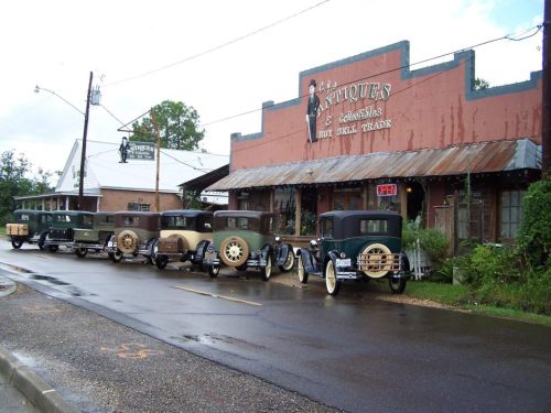 A row of vintage cars parked in front of an antique shop on a rainy street.