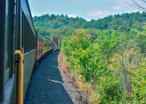 A train travels along a scenic route, surrounded by lush greenery and hills under a clear blue sky.