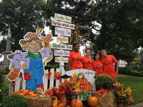 Four people in orange shirts stand by a colorful pumpkin patch sign, surrounded by pumpkins and autumn decorations.