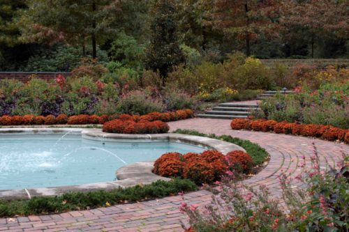 A serene garden with a circular fountain, surrounded by colorful flowers and winding brick pathways.