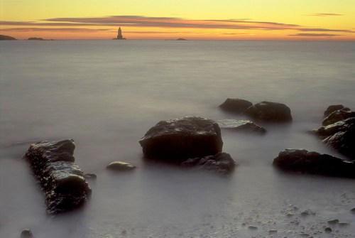 A serene seascape at sunset, featuring smooth rocks in the foreground and a distant lighthouse silhouetted against the sky.