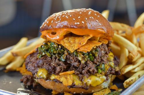 A towering burger with beef, cheese, jalapeños, and nacho chips, served with a side of golden fries.
