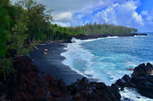 A scenic view of a black sand beach with waves crashing against rocky shores and lush greenery in the background.