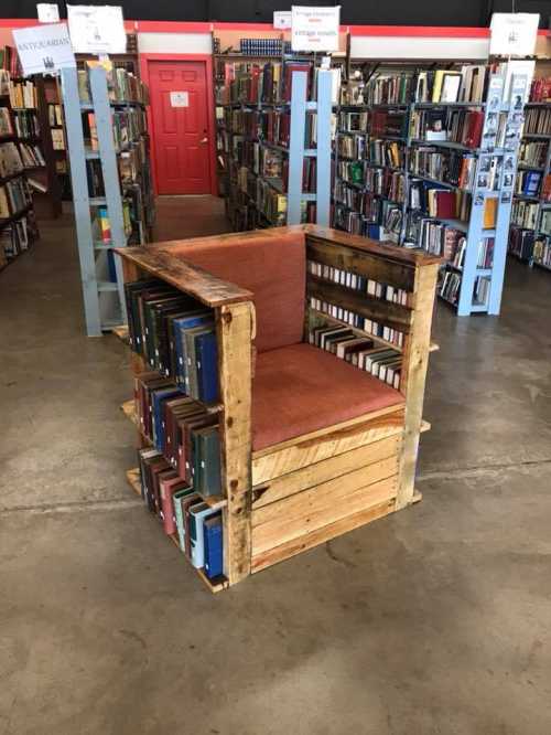 A unique chair made from wooden pallets and books, set in a cozy library with shelves of books in the background.