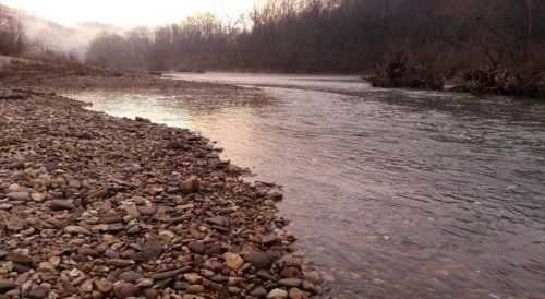 A serene riverbank with smooth stones, reflecting soft morning light and surrounded by misty trees.