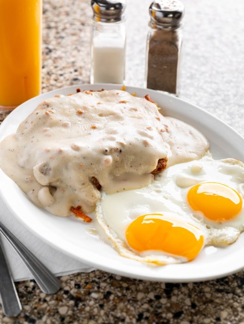 A plate of biscuits smothered in gravy, served with two sunny-side-up eggs, salt, pepper, and a glass of orange juice.