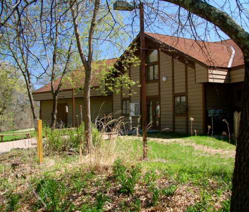 A brown, two-story building surrounded by trees and greenery on a sunny day.