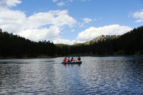 A group of people in life jackets paddles a small boat on a calm lake surrounded by trees and blue skies.