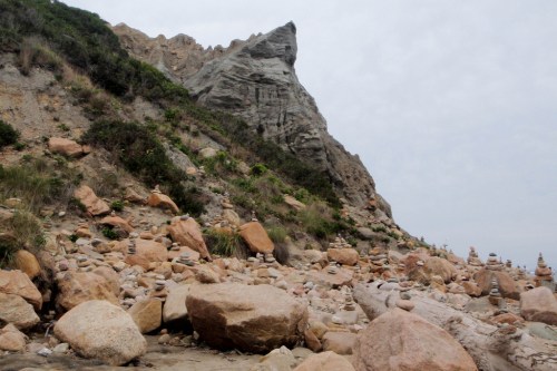 Rocky beach scene with stacked stones and a steep cliff in the background under a cloudy sky.