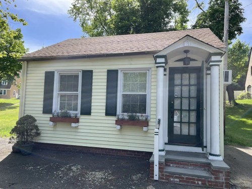 A small yellow house with green shutters, white trim, and a front porch surrounded by greenery.