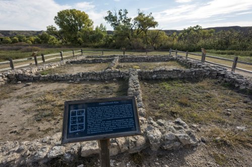 Ruins of a historic structure with a sign explaining its significance, surrounded by greenery and a clear sky.