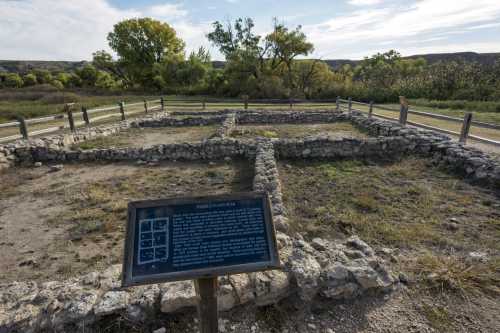 Ruins of a historic structure with a sign explaining its significance, surrounded by greenery and a clear sky.