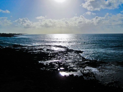 A serene ocean view with sunlight reflecting on the water, rocky shoreline, and a partly cloudy sky.