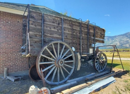 A vintage wooden wagon with large wheels, resting beside a brick building and mountains in the background.