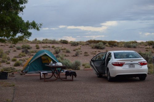 A campsite with a tent, picnic table, and a white car parked nearby under a cloudy sky.