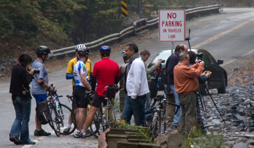 A group of cyclists and photographers gather by a roadside, with a "No Parking" sign visible in the background.